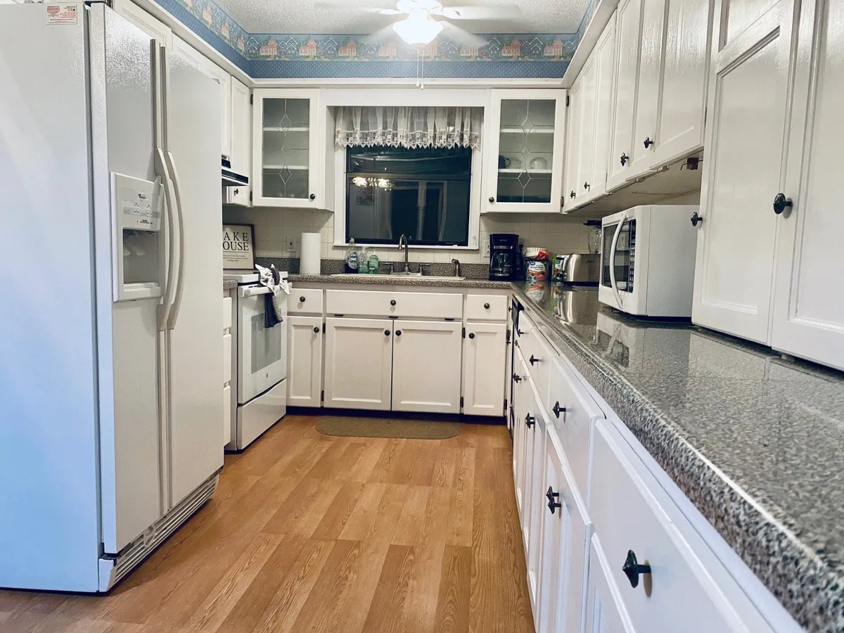 A modern kitchen with granite countertops, white cabinetry, and up-to-date appliances. The room is brightened by a window with lace curtains, creating an organized and welcoming space ideal for pre-stocking essentials and easy meal prep.