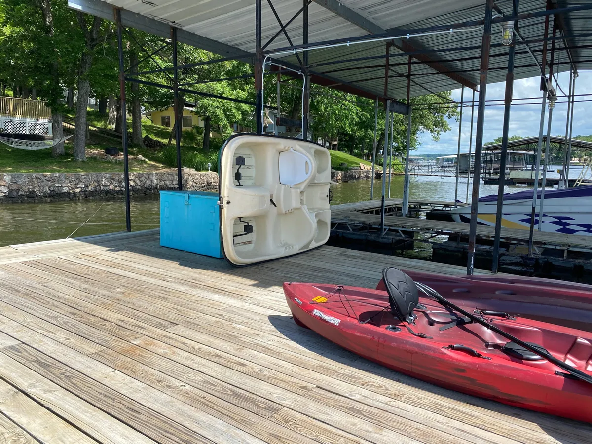 A red kayak rests on the dock, with a blue storage box nearby. The dock extends over calm water surrounded by green trees. The scene suggests easy water access and activities like kayaking and fishing, with the dock providing a clear focal point for lake adventures.