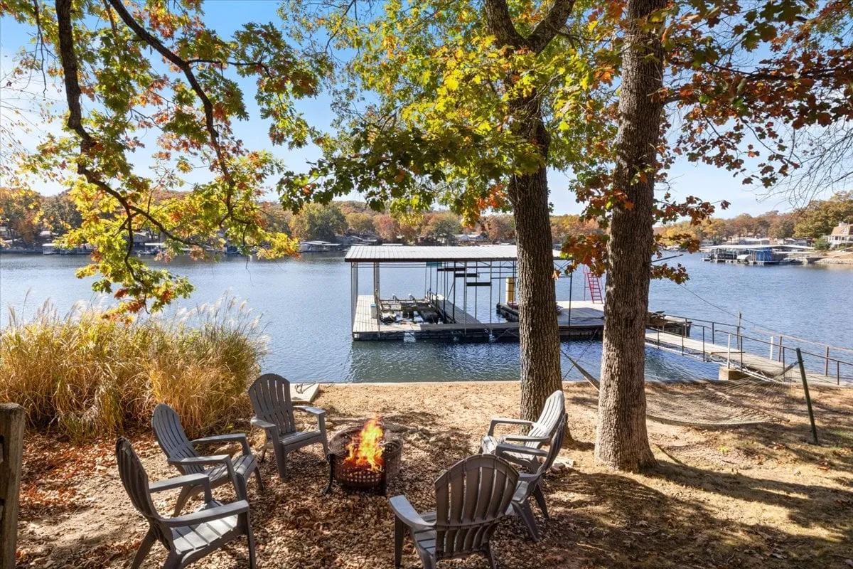 A cozy lakeside fire pit setup with adirondack chairs encircling the fire, set near a dock and the water's edge. The mood is communal and welcoming, ideal for evening gatherings, roasting s'mores, and watching the sunset over the lake.