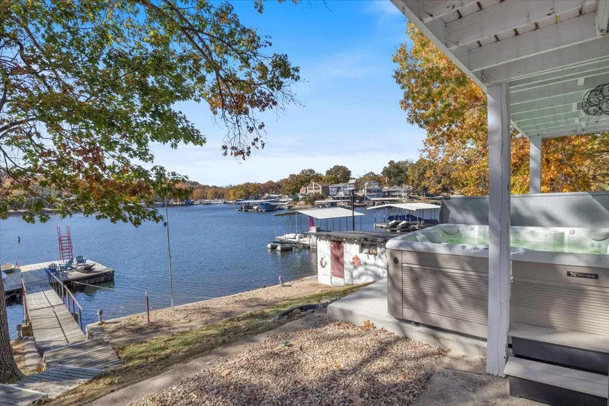 A hot tub sits beside the lake, with a dock extending into the water and trees framing the scene. The landscape is tranquil and inviting, offering a glimpse of relaxation after a day spent enjoying the lake. The firepit and comfortable setting suggest winding down in comfort post-adventure.