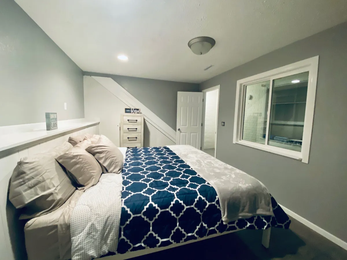 This image presents a cozy bedroom with a patterned bedspread, a nightstand, and a window that filters in soft, natural light. The scene is warm and inviting, suggesting a peaceful environment for relaxation and sleep.