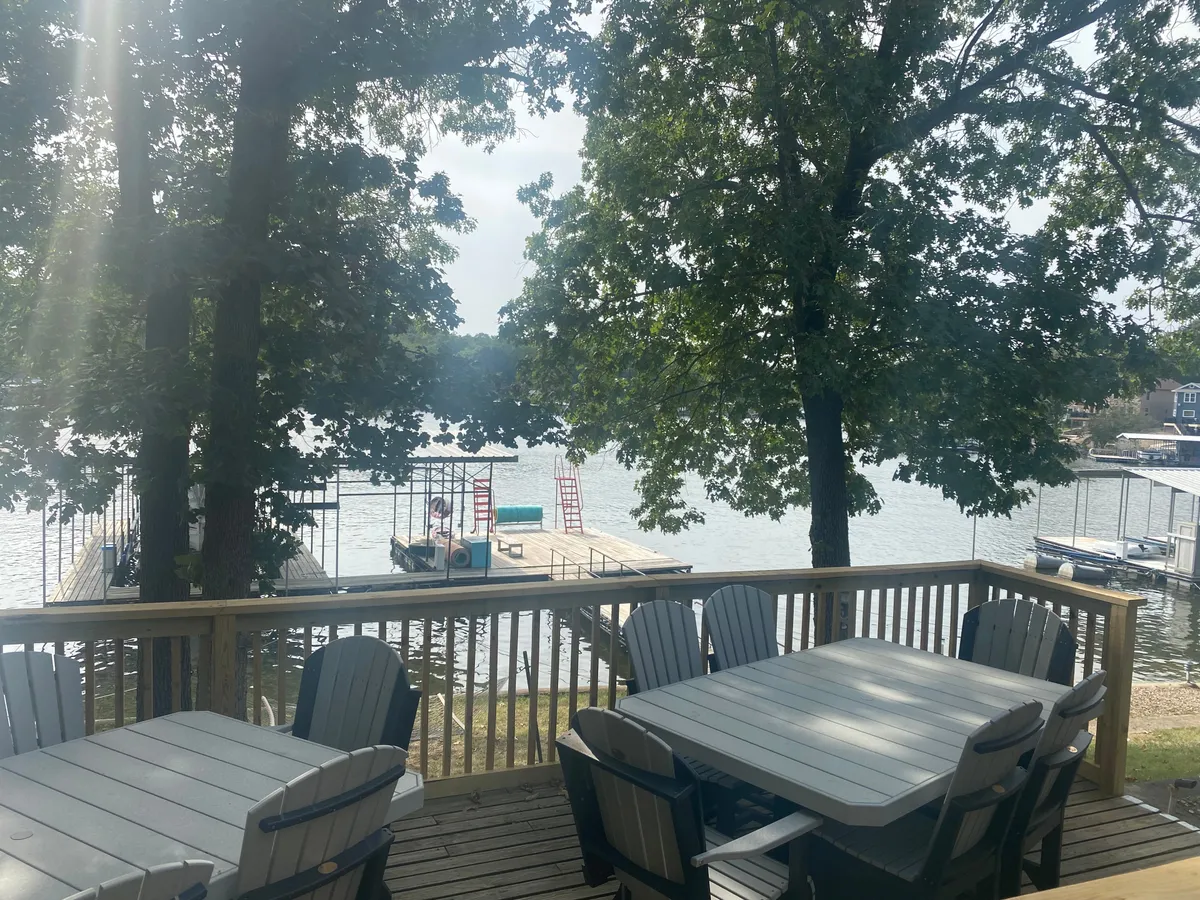 A welcoming dock extends over serene water, surrounded by trees and offering an outdoor seating area. The inviting arrangement hints at boating, fishing, relaxing with friends, and gathering for sunset or morning coffee by the water.