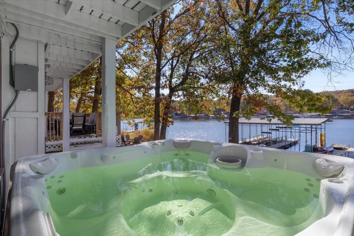 A serene lakeside scene featuring a private hot tub overlooking the water, surrounded by trees and a dock leading out onto the lake. The tranquil atmosphere is amplified by the soft light and the promise of relaxation in nature.