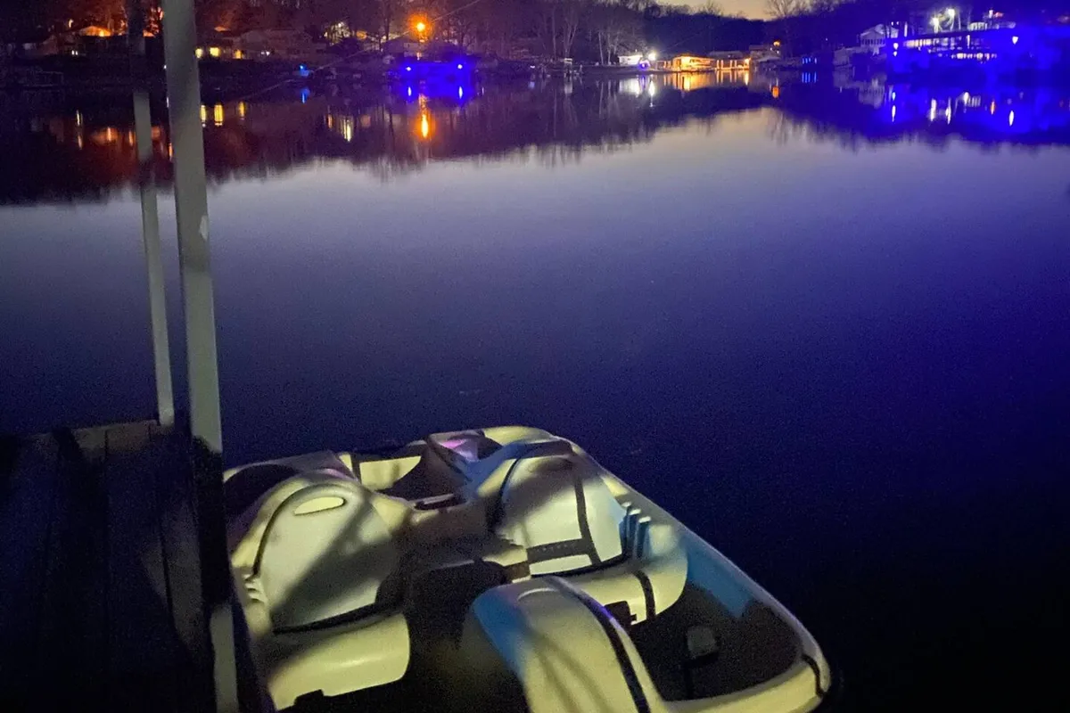 A boat is moored on still, reflective water at sunset or dusk. Warm lights shimmer on the surface, creating a tranquil, almost magical evening scene. The image evokes feelings of peacefulness, with hints of nighttime fishing or stargazing from the water.