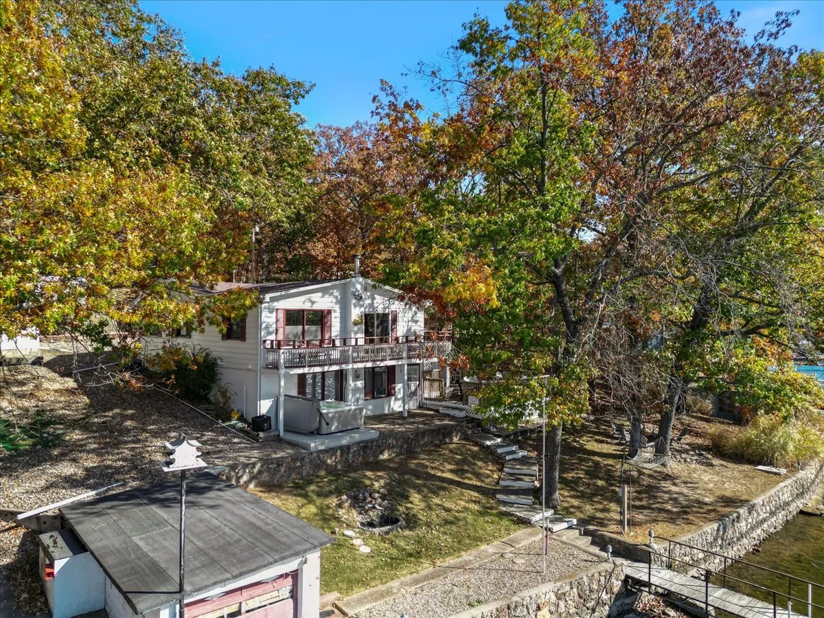 A wide-angle exterior shot of the house with a balcony overlooking the lake, surrounded by lush trees and a landscaped yard, with a clear pathway leading toward the water. The tranquil setting invites both exploration and relaxation.