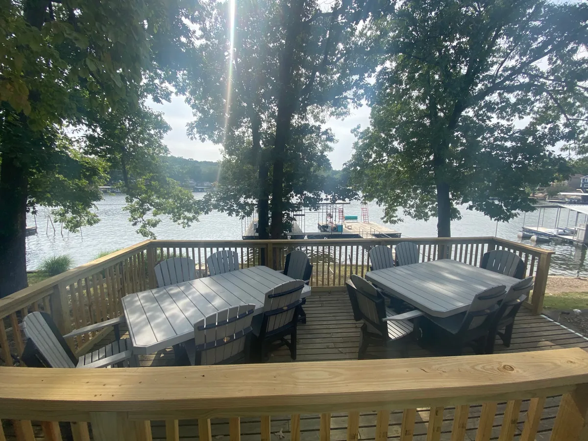 A spacious wooden deck with outdoor seating looks out over the tranquil lake, with trees and nearby docks in view. The deck is staged for relaxing or dining, with ample space for socializing and lingering over morning coffee or sunset drinks while enjoying the water vistas.
