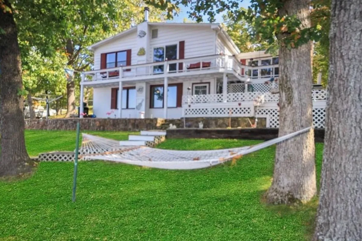 A tranquil exterior view of Buck Creek Cottage surrounded by tall trees, featuring a hammock gently swaying between two trees, an expansive green lawn, and the house in the background. The setting is peaceful, inviting, and perfect for relaxing outdoors after a lake adventure.