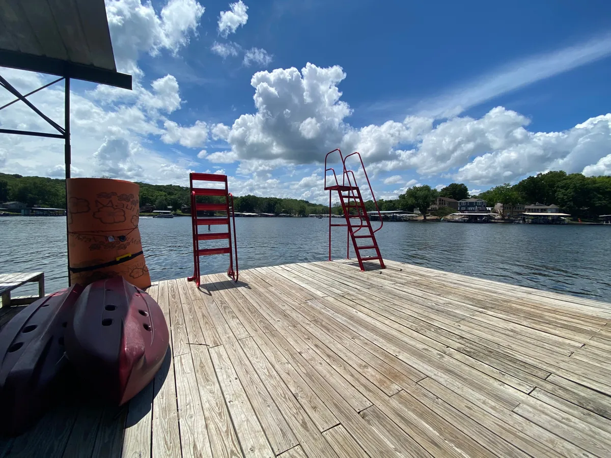 A lively lakeside scene featuring a wooden dock equipped with bright red diving boards and kayaks lined up, all overlooking the shimmering lake. The setup highlights areas for swimming, sunbathing, and water activities, with a sunset glow enhancing the recreational mood.