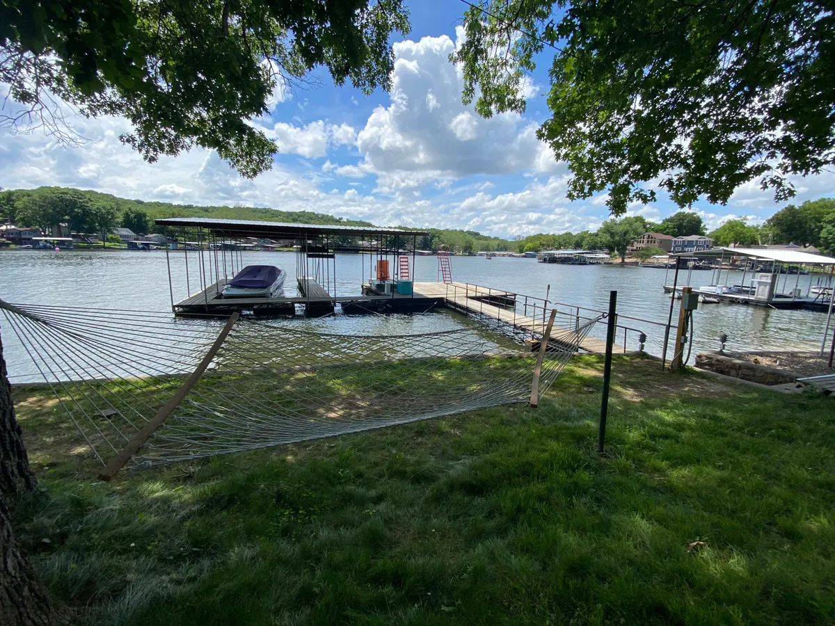 A serene lakeside scene showing a hammock near a dock and the sparkling lake, with boats gently floating in the background. The area is green and shaded, ideal for both people and pets to enjoy nature or relax by the water.